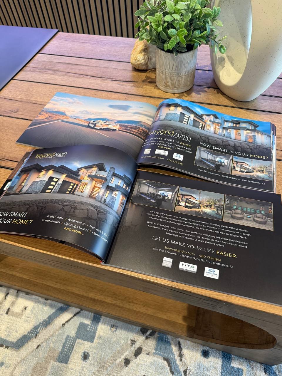 A luxury home and lifestyle magazine open on a wooden coffee table showing a two-page Beyond Audio advertisement — a Scottsdale desert highway on the left, an architectural luxury home and home-theater interior shots on the right, with the Beyond Audio Engineered Environments logo and HTA Certified Estate / Crestron Diamond credentials.