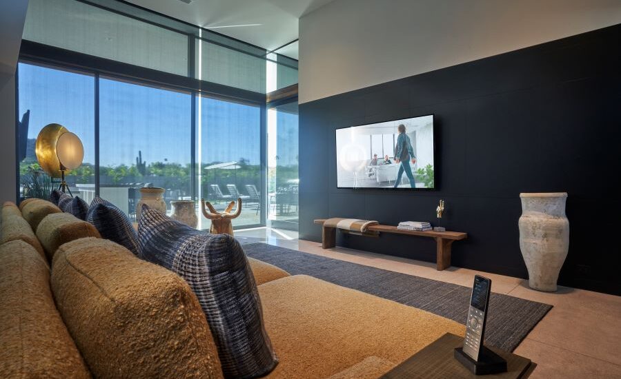 A living room with a couch, TV, Crestron remote, and transparent Crestron shades lowered over floor-to-ceiling windows.