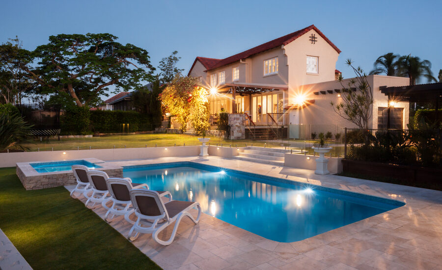 Photo of a beautiful in-ground pool at dusk with lounge chairs beside it with a home featuring interior and exterior lighting in the background.