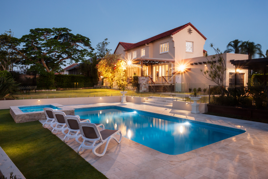 Photo of a beautiful in-ground pool at dusk with lounge chairs beside it with a home featuring interior and exterior lighting in the background.