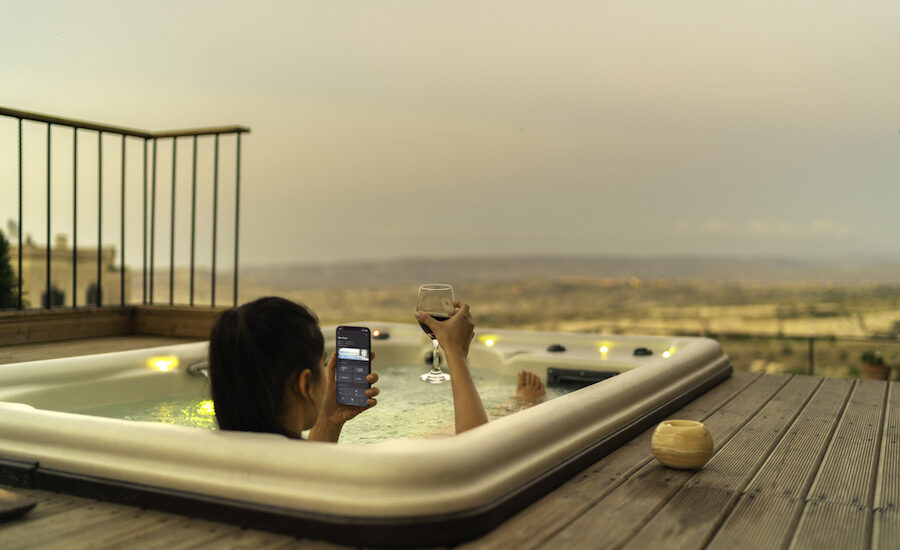 Woman in a hot tub holding a glass of wine while using the Crestron smart home automation app on her phone.
